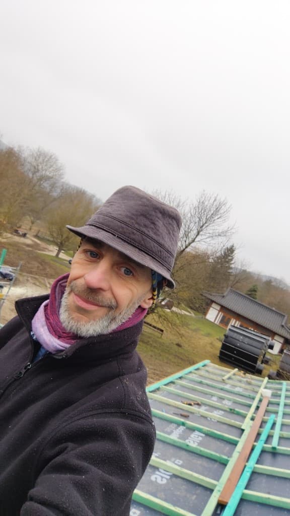 Kerékgyártó László önkéntesként tetőépítés közben a tetőn áll az Eredeti Fény Zen Templomnál: sötét kalapban és fekete polárban, a tetőn zöld lécezés és fekete alátétfólia látszik; háttérben a templom ívelt tetővel, felhős ég alatt.